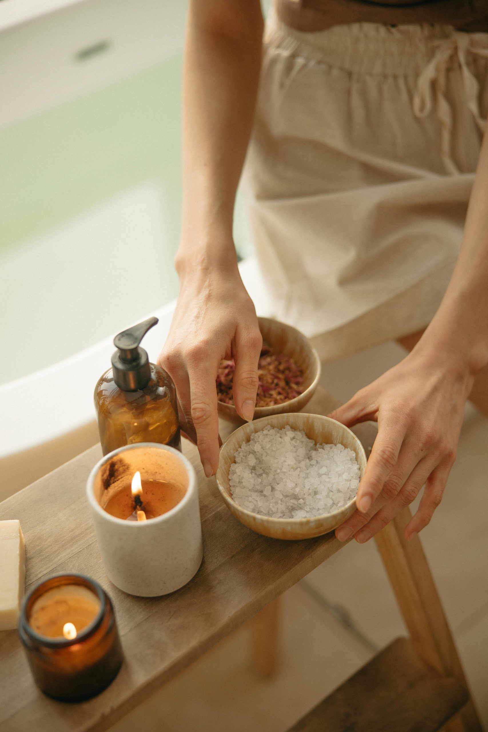 Hands preparing a self-care setup with bath salts, candles, and soap. Serenity Path